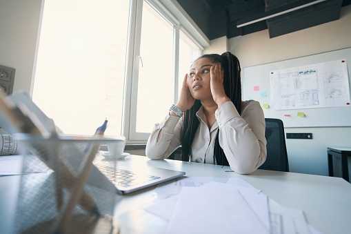 Exhausted female office worker sitting at table and touching her temples with hands