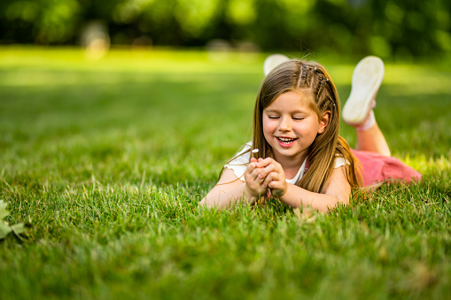 Beautiful little girl laying on grass and picking flowers at the public park on beautiful spring day.