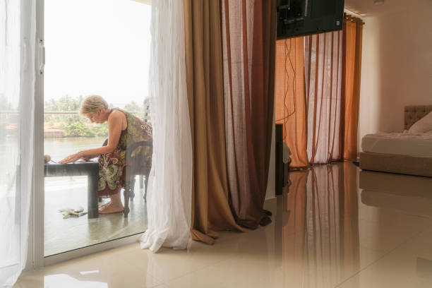 lavoro a distanza in viaggio. donna matura di 55 anni che lavora su un balcone all'aperto in un hotel contemporaneo. la vista da una stanza attraverso il balcone porta aperta. - women mature adult 50 55 years 50s foto e immagini stock