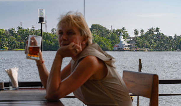 stanca donna caucasica matura di 55 anni seduta in un caffè all'aperto con vista sul fiume bentota, bevendo un bicchiere di birra. messa a fuoco selettiva sullo sfondo con primo piano sfocato. - women mature adult 50 55 years 50s foto e immagini stock