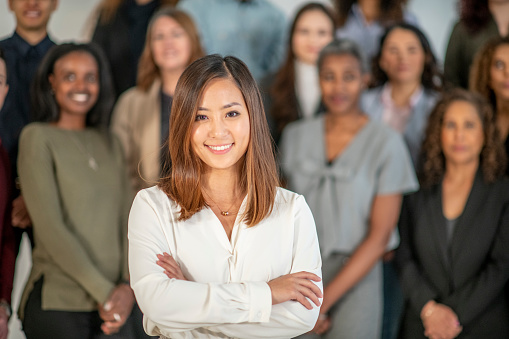 A large group of diverse woman pose together in a studio for a portrait.  They are each dressed semi-casually and are smiling at the camera.  The focus is on a woman of Asian decent in front.