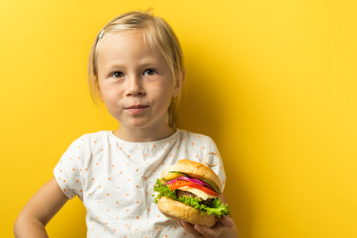 Portrait of cute little caucasian girl with blonde hair enjoying burger on a yellow background. Happy kid eating fast food burger