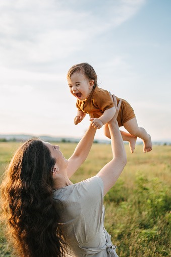 A young mom holding her baby boy up in the sky and they are both laughing while on the beautiful field.