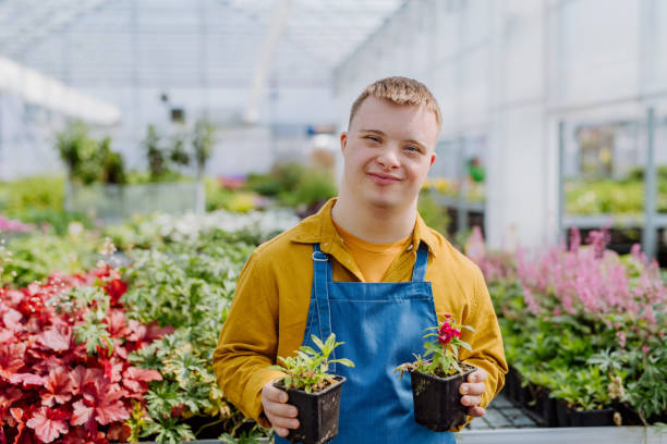 feliz joven empleado con síndrome de down que trabaja en un centro de jardinería, cuidando las flores. - special needs fotografías e imágenes de stock