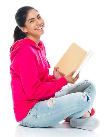 Side view of aged 20-29 years old who is beautiful with black hair generation z female university student sitting in front of white background wearing jeans who is learning and holding textbook