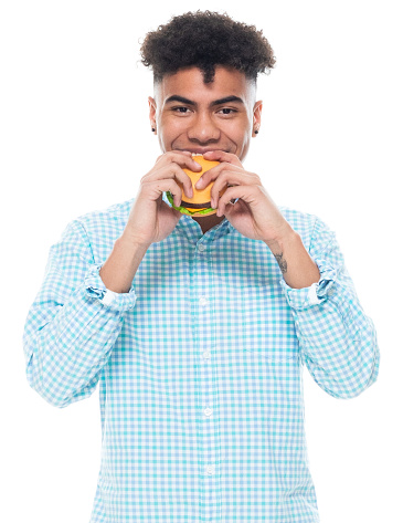 One person of aged 18-19 years old with black hair generation z teenage boys standing in front of white background wearing sports shoe who is hungry and holding hamburger