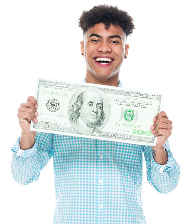 Waist up of aged 18-19 years old with curly hair african ethnicity young male standing in front of white background wearing pants who is smiling and holding us currency