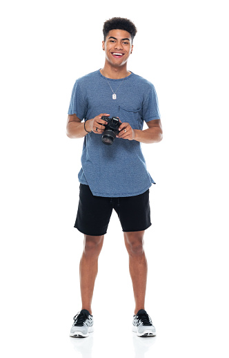 Full length of aged 18-19 years old with black hair african-american ethnicity male photographer standing in front of white background wearing sports shoe who is smiling who is photographing and holding camera