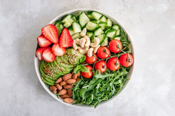 Bowl of side salad composed of arugula, cherry tomatoes, cucumber, strawberries, avocado, almonds and cashews on a minimal background