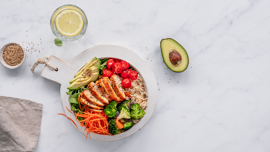 Overhead view of a bowl with grilled chicken, rice, veggies and avocado, next to an avocado half, glass of water with lemon slice, small bowl with sesame seeds and a linen napkin, with copy space on the right side