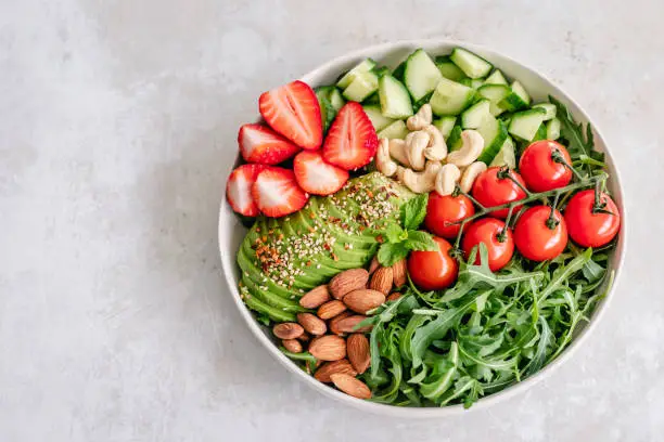 Bowl of side salad composed of arugula, cherry tomatoes, cucumber, strawberries, avocado, almonds and cashews with copy space on the left side