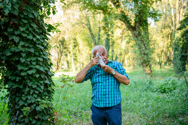 A senior Caucasian man is standing in a forest, wiping his eyes from having an allergic reaction.