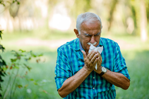 A senior Caucasian man is sneezing in his tissue, while having an allergic reaction in a public park.