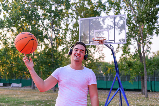 A young Caucasian man wearing headphones is balancing a basketball on his finger with a smile on his face, while standing in a public park.