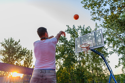 A young Caucasian man is throwing a basketball,, while wearing headphones, with the sun setting behind him.