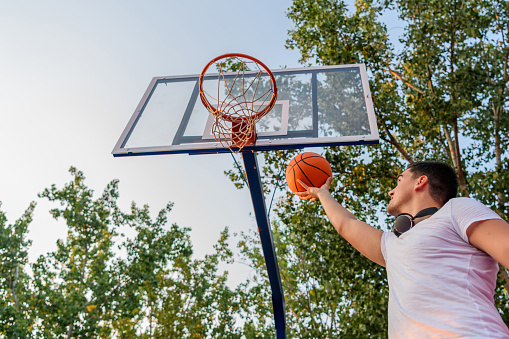 A young Caucasian man wearing headphones around his neck is holding a basketball in his hand, going for the hoop.