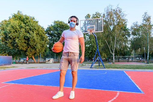 A young Caucasian man is standing on a basketball court holding a ball, while wearing headphones and a protective face mask.