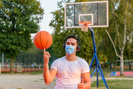 A young Caucasian man is balancing a ball on his finger, while standing on a basketball court, wearing a protective face mask and headphones.