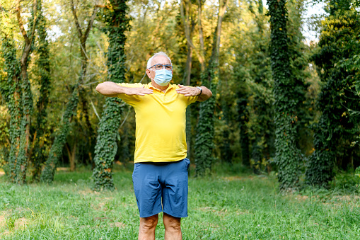 A senior Caucasian man is in a public park, doing his morning excercise, while wearing a protective face mask.