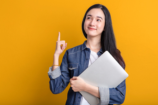 Happy Portrait smiling young asian woman holding laptop isolated on yellow background , Beautiful asian female pointing finger upward and holding notebook computer
