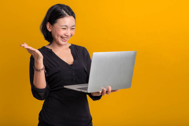 Happy Portrait smiling young asian woman holding laptop isolated on yellow background , Beautiful asian female pointing finger upward and holding notebook computer stock photo