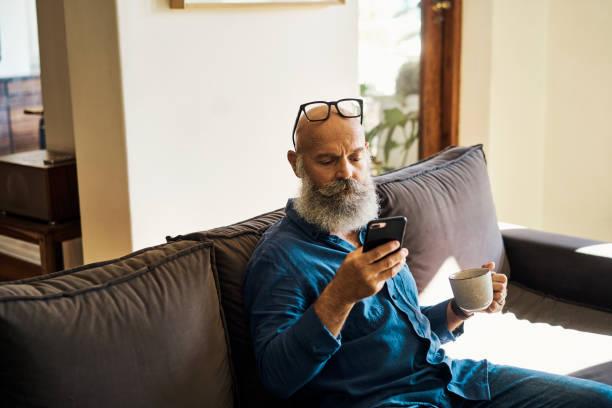 istock Senior man using a phone searching the internet while drinking a cup of tea on couch at home. Mature male relaxing and scrolling on social media online on the sofa. Retired guy typing a text message 1407638069
