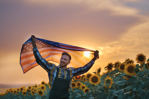 Proud farmer running and waving the American flag in a sunflower field during sunset. 4th of July concept.