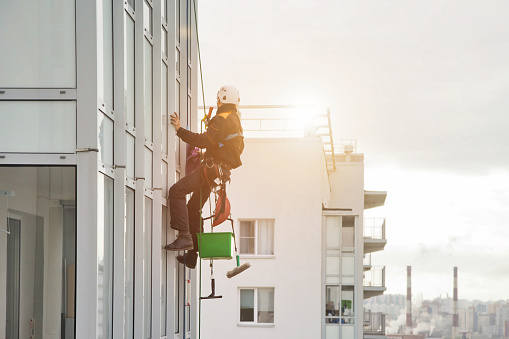 Industrial mountaineering worker in uniform hangs over residential facade building, washing exterior glazing. Rope access laborer hangs on wall of house. Concept of industry urban works. Copy space