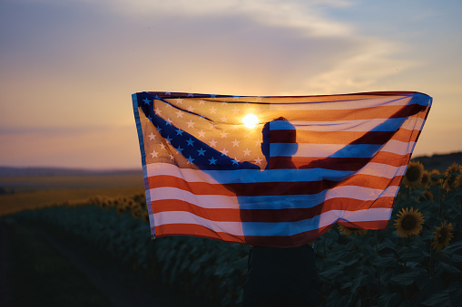 Young man holding American flag on back while standing in sunflower field. Looking at the sunset