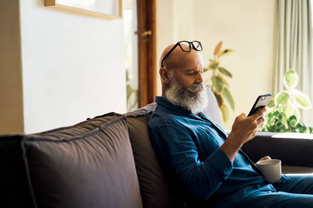 istock Mature man texting on phone, browsing social media, searching internet and relaxing alone on home living room sofa. Drinking cup of coffee or tea while enjoying online videos. Downloading dating app 1407638371