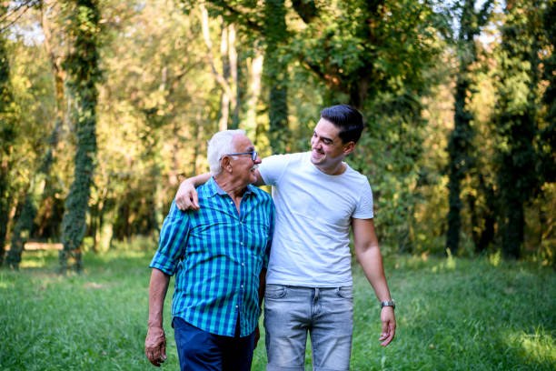 A young Caucasian man is embracing his elderly grandpa, while taking a walk through the park together.
