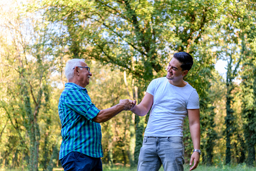 A young Caucasian man and his grandpa are cheerfully bumping fists, while spending time in the park totgether.