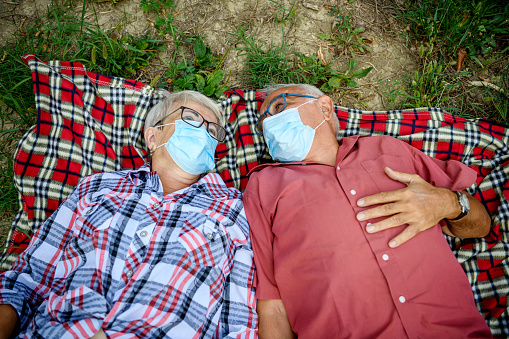 A senior Caucasian couple is lying down on their picnic blanket, wearing protective face masks.