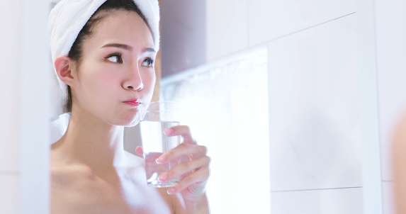 asian Young woman with perfect fresh skin and white towel on head - she is rinsing mouth by glass of water in the bathroom
