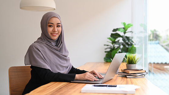 Elegant Muslim woman wearing hijab using computer laptop in bright modern office.