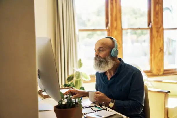 Photo of Senior man wearing headphones to listen to music while working on a desktop computer at home. Retired male browsing internet online and enjoying favourite songs. Mature hipster guy watching videos