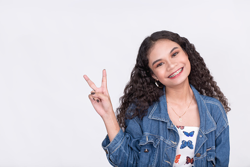 A beautiful and lovely young asian lady with natural curly hair makes a peace sign. Wearing a blue denim jacket. Isolated on a white background.