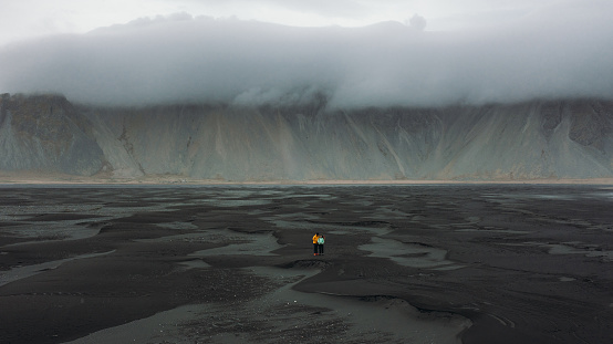 Drone photo of a young couple of travelers enjoying the cloudy morning walking at the black sand remote Stokksnes beach with mounain view at East Iceland
