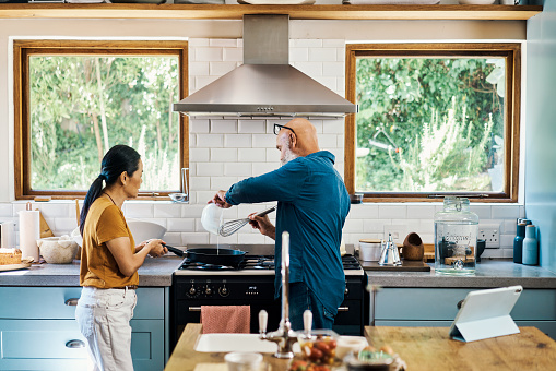 Mature interracial couple using a digital tablet to cook breakfast and prepare pancakes in a home kitchen for romantic brunch. Loving husband and wife following online fried recipe for a healthy meal