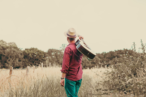 A man walks alone on a rural path surrounded by golden summer meadows and tall grass. He carries an acoustic guitar, leather bag and he wears a red check shirt, panama hat and sunglasses. Room for copy space.