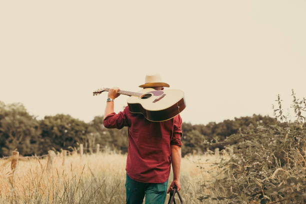 A man walks alone on a rural path surrounded by golden summer meadows and tall grass. He carries an acoustic guitar, leather bag and he wears a red check shirt, panama hat and sunglasses. Room for copy space.