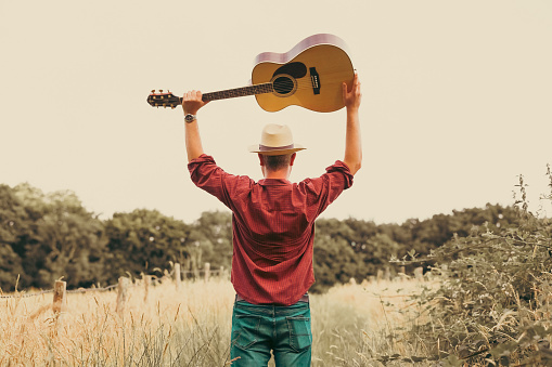 A man walks alone on a rural path surrounded by golden summer meadows and tall grass. He carries an acoustic guitar, leather bag and he wears a red check shirt, panama hat and sunglasses. Room for copy space.