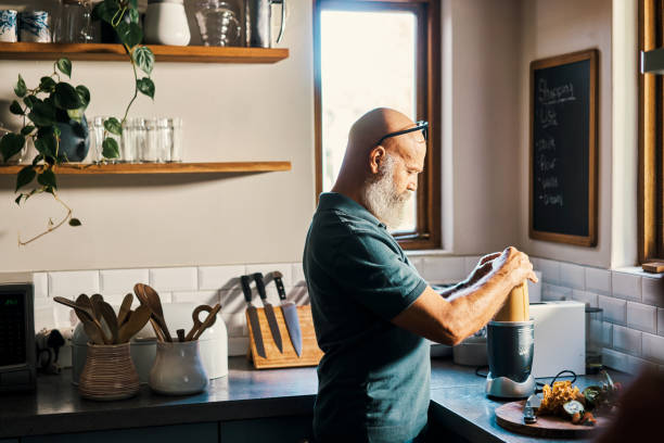Profile of older, senior man cooking in a kitchen with a bullet blender inside, making dinner. Casual mature bald male with grey beard making soup or a vegetable smoothie in a modern bright home
