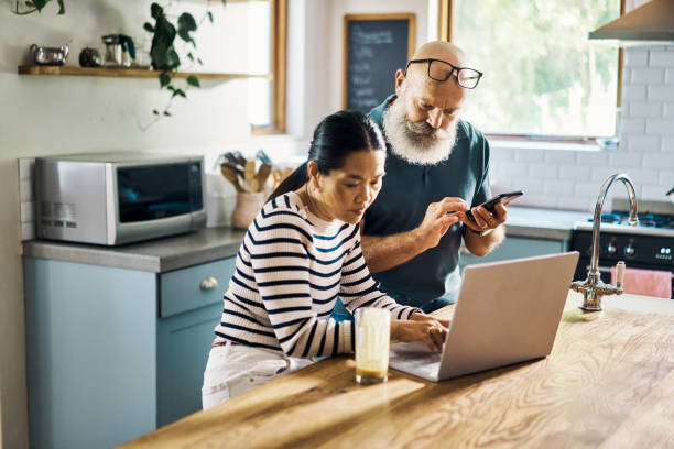 una pareja madura que usa una computadora portátil y un teléfono en la cocina de casa. marido y mujer navegando en línea usando aplicaciones de redes sociales con sus dispositivos inalámbricos en casa. hombre y mujer navegando, comprando en línea - standing digital tablet couple love fotografías e imágenes de stock