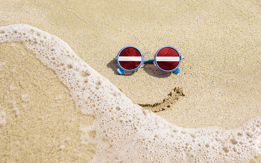 Sunglasses with flag of Latvia on a sandy beach. Nearby is a sea lightning and a painted smile. The concept of a successful vacation in the resorts of
Latvia.