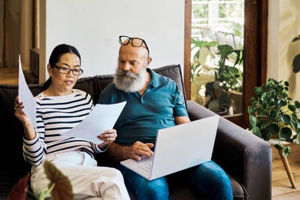 pareja interracial mayor usando una computadora portátil y yendo a los documentos de jubilación en línea en el sofá de casa. esposo y esposa maduros hablando y leyendo documentos mientras usan una pc en el sofá del salón - retirement 401k finance pension fotografías e imágenes de stock