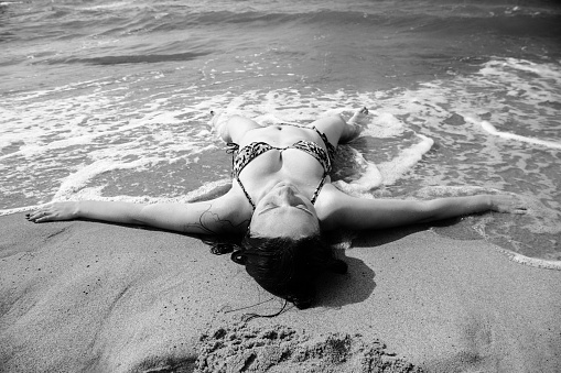 single happy young brunette woman in bikini lying at sea surf with splashes, monochrome