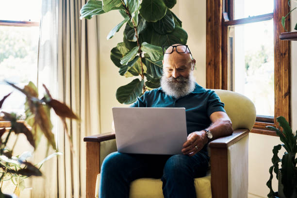 istock Mature hipster male relaxing and scrolling on social media on a pc. Retired guy watching series. Senior man working on a laptop and searching the internet online while sitting on a chair at home. 1407678024
