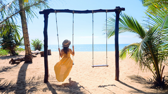 Woman tourist swinging on swing, sandy beach with palm trees and blue sea on background. Tropical vacation. Girl in yellow dress on summer holidays, fun on sunny coastal, good mood, romantic.