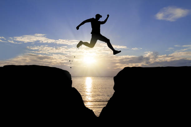 silueta del hombre saltando sobre el acantilado sobre el fondo del amanecer, logro del concepto de negocio - hurdle fotografías e imágenes de stock